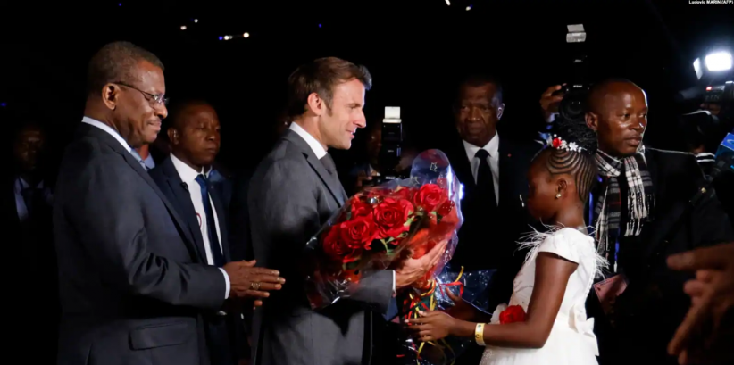 Une jeune fille offre des fleurs au président français Emmanuel Macron à son arrivée à Yaoundé, où il a été accueilli par le Premier ministre Joseph Dion Ngute (à g.), le 25 juillet 2022.